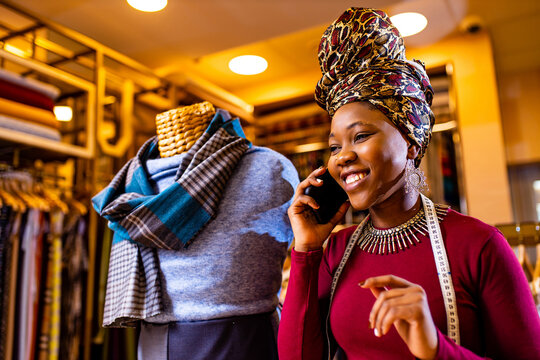 Tanzanian Woman With Snake Print Turban Over Hear Working In Fabrics Shop Calling To Client By Smartphone