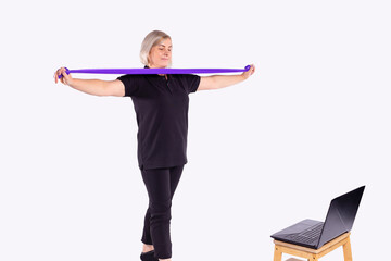 Front view of senior caucasian woman at home watching online video on laptop computer for training, using violet rubber resistance band for workout on gray background. Health fitness concept copyspace