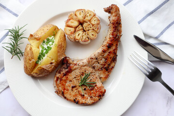 Grilled pork chop steak with baked potato and roasted garlic in white plate on marble background. Close up view