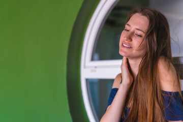 A smiling young pretty redhead Caucasian woman sitting in a blue dress next to a white sale of a green house, copy space and paste
