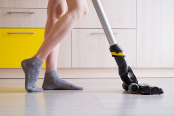 The upright vacuum cleaner cleans the laminate floor in the kitchen. Women's legs. Gray and yellow tints. The concept of cleaning the house, keeping it clean.