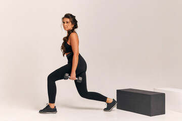 Fototapeta premium Athletic young woman in a black suit lunges forward with dumbbells on a white background. crossfit. fitness squats healthy lifestyle