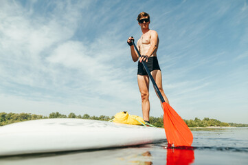 Obraz premium Athletic man in sunglasses swims on a sup board on the pond, paddles with a serious face and looks at the camera. Active summer vacation on the pond.
