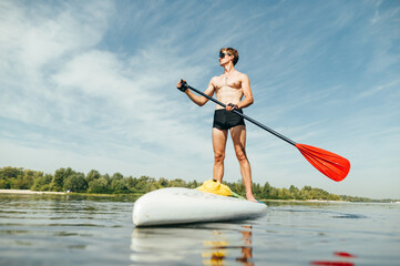 Athletic muscular man with a deaf torso stands on a sup board with an oar in his hand and looks away with a serious face.