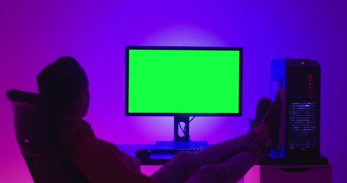 Young man freelancer resting on a chair, drinking coffee and looking at a computer monitor with a green screen chroma key, indoors at night in room filled with neon light.