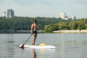 Athlete with an athletic body with an oar in his hands stands on a sup board and looks to the side against the backdrop of a beautiful landscape with a river and park. Surfing on the sup board