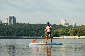 Muscular young man stands on sup board on water and paddles on background of beautiful landscape. Fitness on sup board