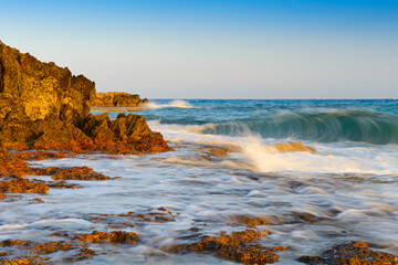 Turquoise sea with waves and splashes and a rocky orange beach on a summer evening