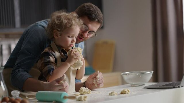 man and his little son are having fun at kitchen, cooking and playing with dough, learning to cook by internet