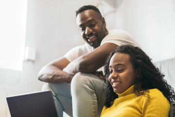 african american couple smiling and watching laptop and laughing at home. sitting on the sofa and the floor