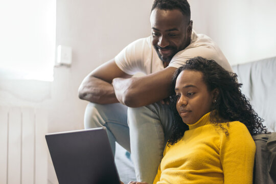 African American Couple Watching Laptop And Laughing At Home. Sitting On The Sofa And The Floor