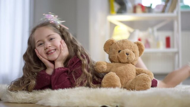 Playful beautiful little girl lying on carpet with teddy bear smiling and looking at camera. Portrait of joyful pretty Caucasian child posing at home with toy indoors. Happiness and leisure
