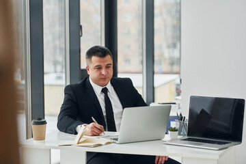 Man in suit and tie sits by table with laptop and works in the office