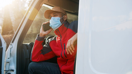 Man with medical mask sitting on the drivers seat of the van and having a phone call. High quality photo
