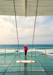 Mujer con chaqueta roja mirando el mar desde un mirador de metal y cristal