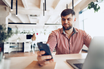 Young man in casual shirt using smartphone in office while sitting at his desk