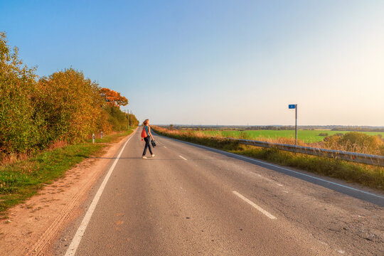 Solo Journey Concept. Back View Of Woman With Backpack Crosses The Autumn Country Road, Hiking Trip