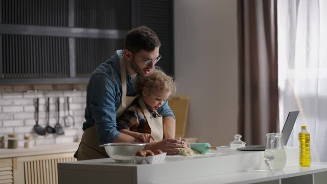 dad and toddler son are cooking by internet recipe, looking on laptop and making dough on kitchen table, father and baby boy