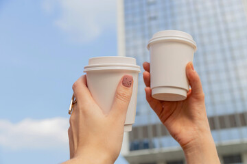 Two white cups with coffee to go in female hands against the background of city buildings and the sky. High quality photo