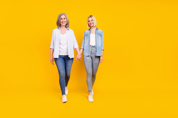 Full length body size photo mother and daughter wearing denim outfits holding hands going forward isolated vibrant yellow color background