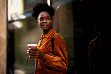 Young african woman enjoying outdoors. Beautiful woman with curly hair drinking coffee while walking around town.