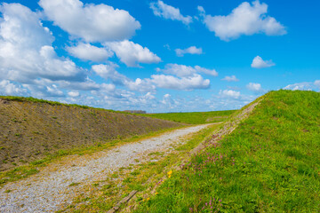 Fototapeta na wymiar Dikes in a green grassy field below a blue cloudy sky in sunlight in spring, Almere, Flevoland, The Netherlands, April 13, 2021