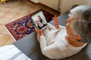 Mature woman sitting on sofa at home greets young doctor in remote video call on tablet for a medical consultation - Elderly patient in quarantine at house - Psychologist during psychotherapy session