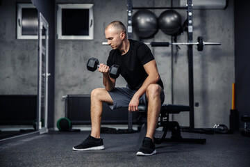 Fototapeta na wymiar Pumping arm and shoulder muscles. A man in a black T-shirt lifts a dumbbell with one hand while sitting in the gym. Strengthening the triceps and biceps. Sports lifestyle and fitness challenge