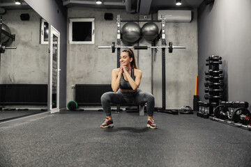 Fototapeta na wymiar Squatting and burning the muscles of the buttocks and legs. Portrait of a hot woman in sportswear and good physical shape doing squats in an isolated indoor gym. Strength and motivation, fitness goal