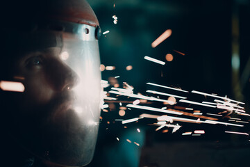 Man welder grinder in transparent protective mask with flying sparks in darkness.