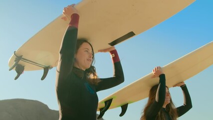 Handheld shot of two women in wetsuit outfits carry surfboards towards ocean. Cinematic sun flares camera spin around beautiful inspiring surf athletes on sunny day. Summer wanderlust vibes
