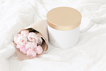 Gold gift box on the bed. Bouquet of white peonies in craft packaging. Scandivanavian white interior.