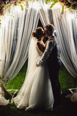 Beautiful newlyweds hugging against the backdrop of flowers and green arch with lamps and candles at night. Film noise.