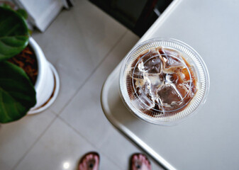 Top view of an iced coffee cup on the table's corner, trees, and human feet