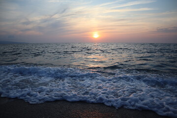 Fototapeta premium Scenic sunset by the sea in pastel colors, Calabria, Italy. Seascape sun on the horizon reflected in waves and sea foam at sunset on a summer evening