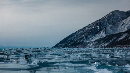 Transparent shiny ice floes are scattered over the surface of the frozen lake to the horizon. Against the background of the sky, there is a wooded and snow-covered mountain. Pastel shades. Baikal