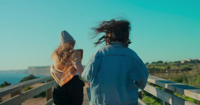 Handheld authentic and real POV shot of friends running together at sunset wooden boardwalk. Happy teenage freedom vibes, summertime lifestyle of travel wanderlust. Female support and friendship