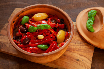 Roasted bell peppers with potatoes, olives, garlic, basil leaves and tomato sauce in clay pot. Italian cuisine peperonata dish.