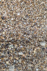 Top view of round flat colorful pebbles, sand and seashells, wet by sea waves. Natural marine pattern, half of the vertical frame blurred under water. Fresh summer holiday inspiration, relaxation mood