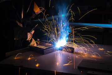 A man welder with construction gloves and a welding mask is welded with a welding machine metal in workshop.