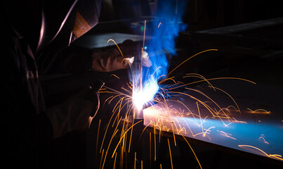 A man welder with construction gloves and a welding mask is welded with a welding machine metal.