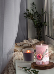 Beautiful decorated candles by the window on a sill with a blanket, glasses and an open book. Cozy atmosphere in the house.