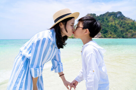 Happy Asia boy kiss mother and standing at the beach in Thailand on sunny day with blue sky. Smile together and enjoy on summer lifestyle travel holiday vacation. Positive human emotions and love