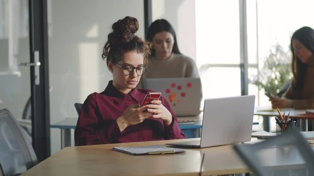 Young attractive businesswoman sitting at workplace in office and surfing the Internet on smartphone