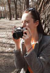 A young girl, a brunette in a gray coat sits near a large tree in the park, holds an old film camera in her hand and looks to the side