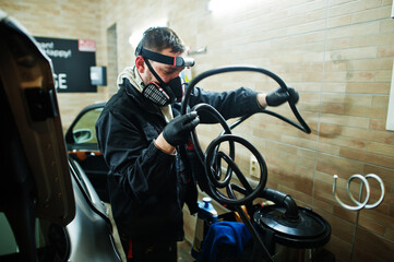 Man in uniform and respirator, worker of car wash center, cleaning car interior with hot steam cleaner. Car detailing concept.