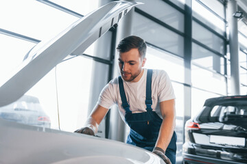 Quality service. Young man in white shirt and blue uniform repairs automobile