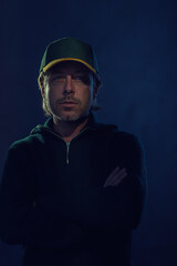 Fototapeta premium Shaded dark portrait of a blond man with stubble beard and baseball cap in a dark green wool sweater in front of a dark blue wall.