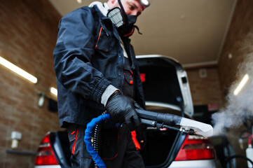 Naklejka premium Man in uniform and respirator, worker of car wash center, cleaning car interior with hot steam cleaner. Car detailing concept.