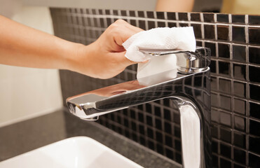 Closeup of hand using tissue wipes cleaning the water faucet before using it to prevent coronavirus infection, dust and germs. Selective focus on wipes.